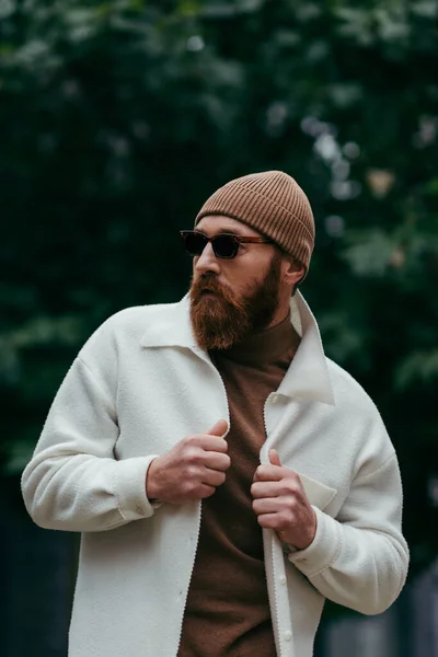 Bearded man in stylish sunglasses and beanie hat adjusting white shirt jacket near green leaves — Stock Photo