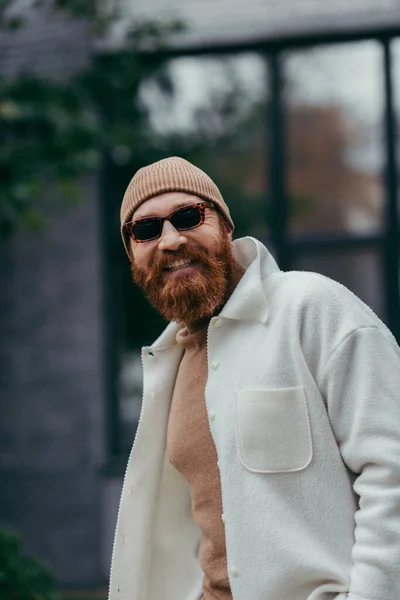 Happy and bearded hipster in stylish sunglasses and beanie hat smiling outside — Stock Photo