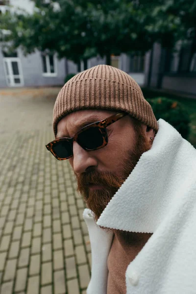 Uomo sospetto con la barba in occhiali da sole alla moda e cappello beanie guardando la fotocamera — Foto stock