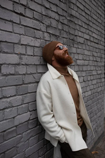 Cheerful and bearded man in beanie hat and sunglasses leaning on brick wall — Stock Photo