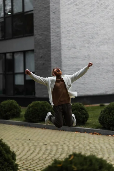 Aufgeregter Mann in trendiger Sonnenbrille und Hemdjacke springt in der Nähe moderner Gebäude — Stockfoto