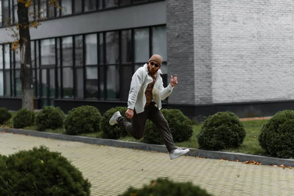 Vollbärtiger Mann mit Hut und trendiger Sonnenbrille springt in der Nähe moderner Gebäude — Stockfoto