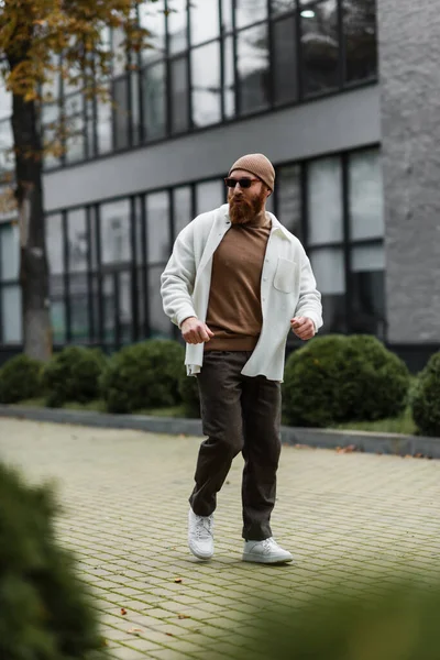 Full length of bearded man in beanie hat and stylish sunglasses walking in shirt jacket near modern building — Stock Photo