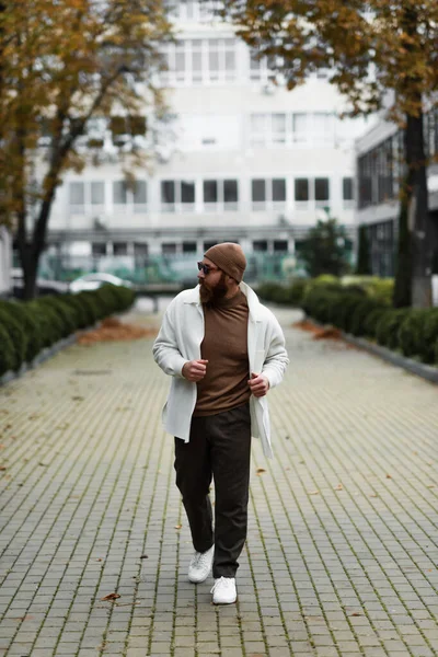 Full length of bearded man in beanie hat and trendy sunglasses walking in shirt jacket near building — Stock Photo