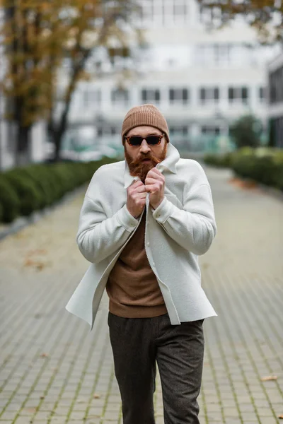Бородатый мужчина в шапочке и модных солнцезащитных очках чувствует холод во время прогулки на улице — стоковое фото