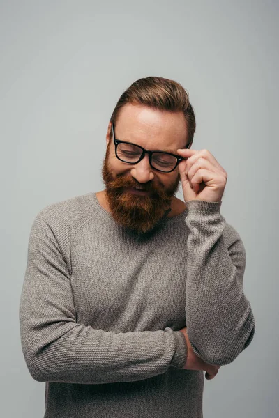 Bearded man in long sleeve smiling while adjusting eyeglasses isolated on grey — Stock Photo