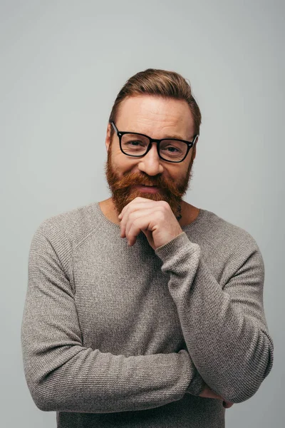 Bearded man in long sleeve and eyeglasses smiling while looking at camera isolated on grey — Stock Photo