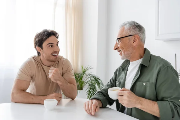 Взволнованный парень жестикулирует, разговаривая с улыбающимся папой за чашкой кофе на кухне — стоковое фото