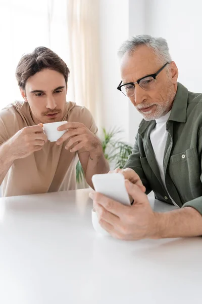 Senior man in eyeglasses showing smartphone to young son drinking coffee in kitchen — Stock Photo