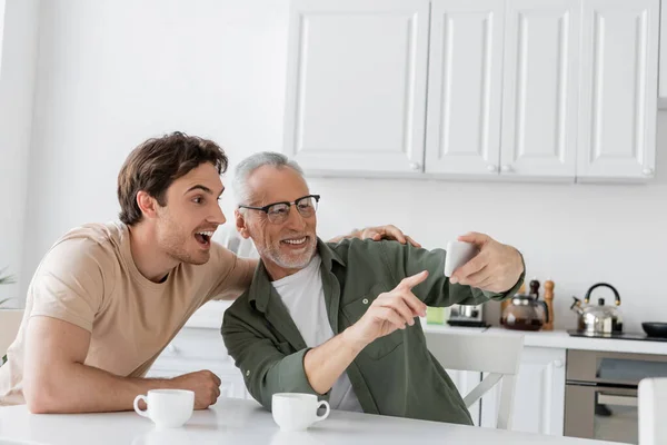 Smiling grey haired man pointing at mobile phone near amazed son and coffee cups on kitchen table — Stock Photo