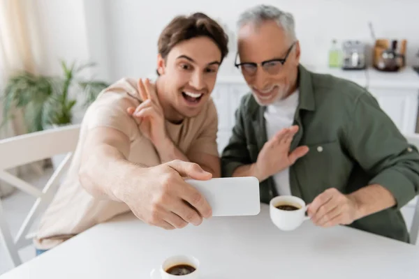 Cheerful young man showing victory sign while taking selfie with mature dad holding coffee cup and waving hand — Stock Photo