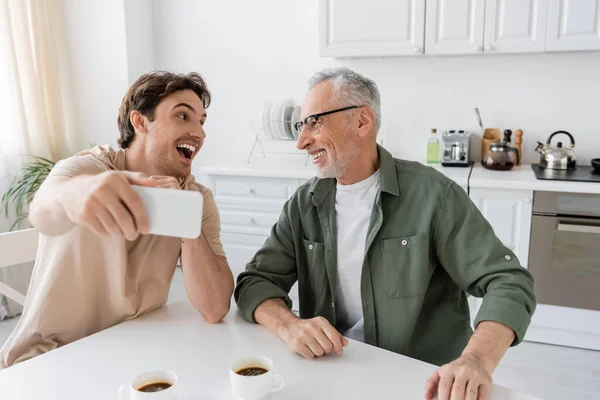 Joven hombre excitado con teléfono móvil tomar selfie con padre sonriente cerca de tazas de café en la cocina - foto de stock