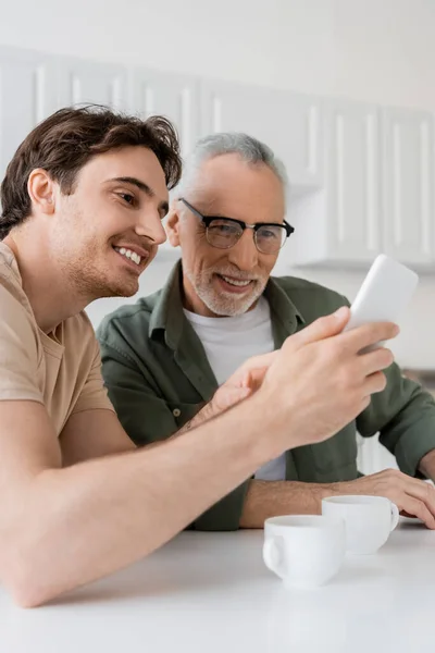 Улыбающийся мужчина показывает мобильный телефон улыбающемуся папе возле кофейных чашек на кухне — стоковое фото
