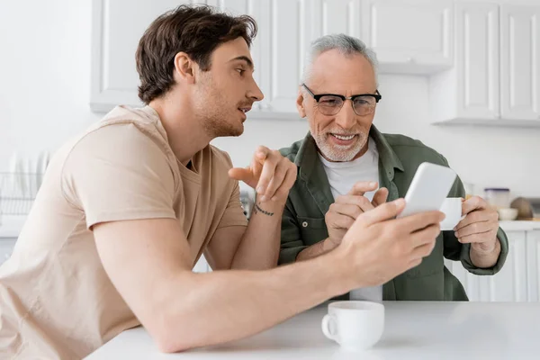 Junger Mann zeigt fröhlichem Vater Handy, der mit Kaffeetasse in Küche sitzt — Stockfoto