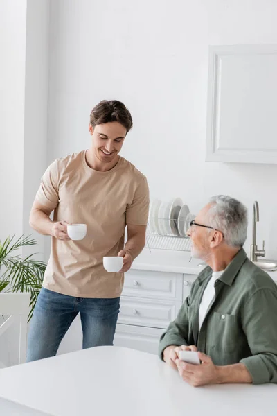 Young man smiling while holding coffee cups near dad sitting with smartphone in kitchen — Stock Photo