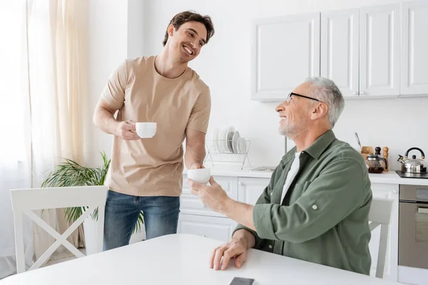 Young and happy guy giving coffee cup to smiling grey haired father sitting in kitchen — Stock Photo