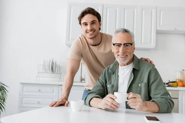 Homem maduro feliz com xícara de café e filho jovem feliz olhando para a câmera na cozinha — Fotografia de Stock