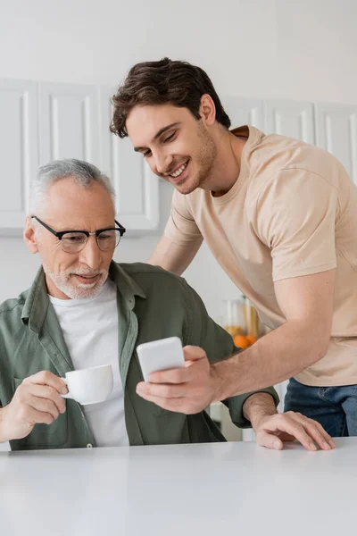 Young man showing mobile phone to father drinking morning coffee in kitchen — Stock Photo