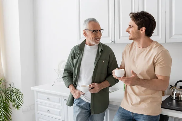 Smiling mature man holding coffee cup while standing with hand in pocket and listening to son in kitchen — Stock Photo