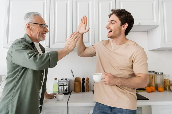 Smiling man giving high five to excited son with coffee cup in kitchen — Stock Photo