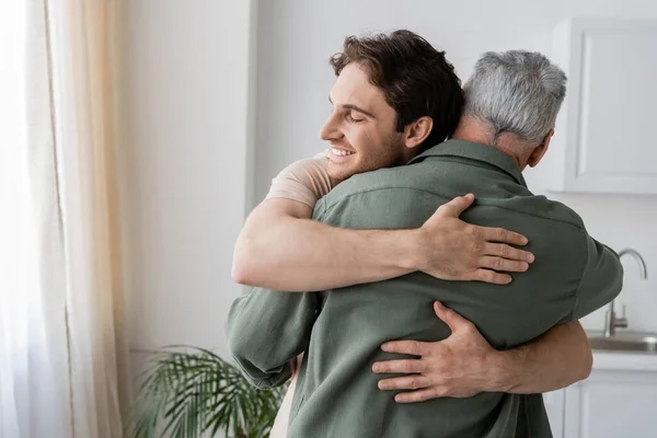 Glücklicher junger Mann mit geschlossenen Augen, der den reifen Vater in der Küche umarmt — Stockfoto