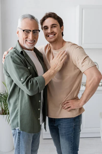 Young and smiling man hugging shoulders of happy father touching his chest and smiling at camera — Stock Photo