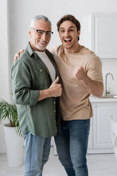 Cheerful father and son showing like and success gestures while looking at camera in kitchen — Stock Photo