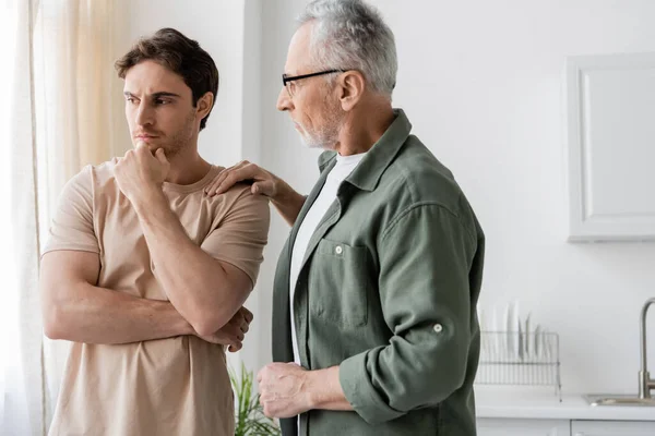 Thoughtful and upset man holding hand near face while father calming him in kitchen — Stock Photo