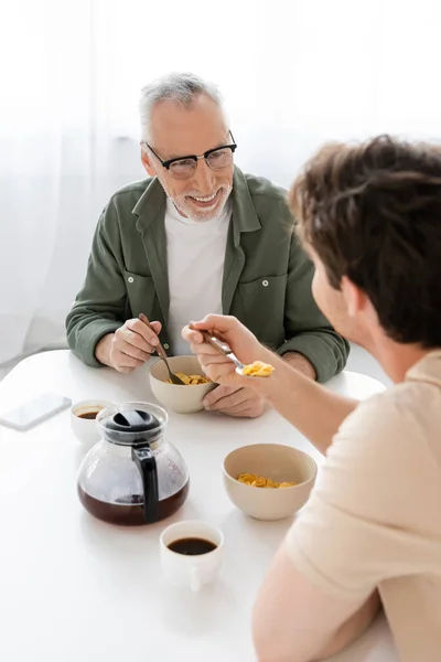 Sonriente hombre en gafas mirando a su hijo joven comiendo hojuelas de maíz para el desayuno - foto de stock