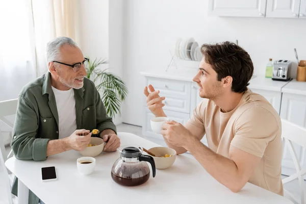Young man with coffee cup gesturing and talking to smiling dad near bowls with corn flakes in kitchen — Stock Photo