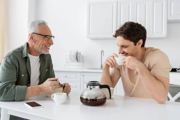 Uomo sorridente guardando il figlio che tiene in mano la tazza di caffè e ride con gli occhi chiusi durante la colazione — Foto stock