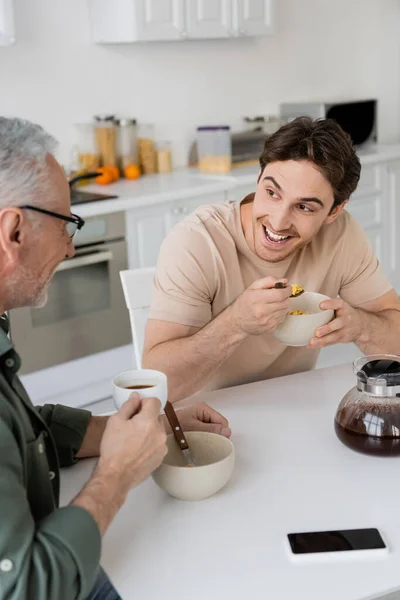Hombre sorprendido escuchando a padre mientras que come hojuelas de maíz cerca de la cafetera en la cocina - foto de stock