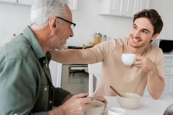 Cheerful young man holding coffee cup and touching shoulder of dad while talking to him during breakfast — Stock Photo