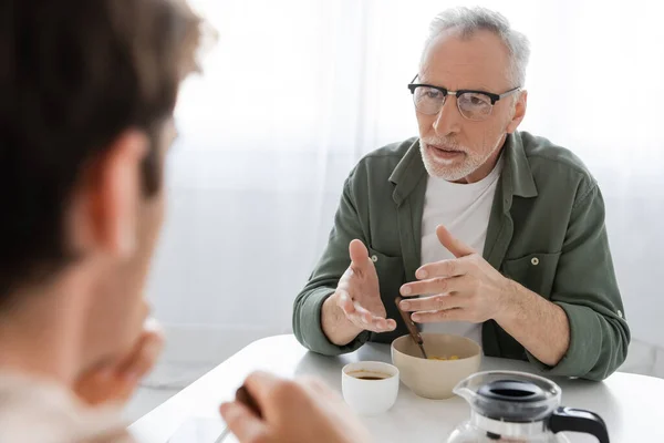 Senior man in eyeglasses gesturing during conversation with son having breakfast in kitchen — Stock Photo