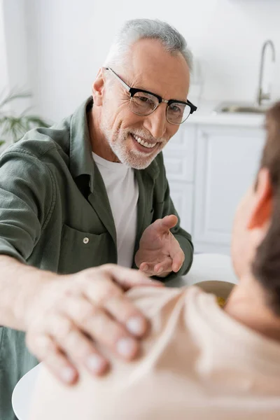 Smiling man in eyeglasses pointing with hand and touching shoulder of blurred son during conversation in kitchen — Stock Photo