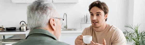 Joven con taza de café gesto y hablar con el padre borroso en la cocina, pancarta — Stock Photo