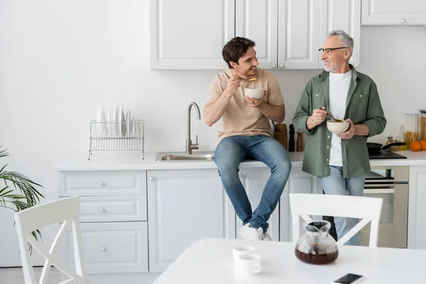 Young man sitting on kitchen worktop while having breakfast and talking to smiling dad — Stock Photo