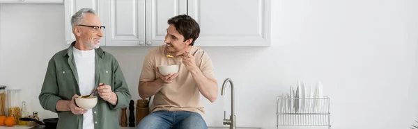 Cheerful young man having breakfast and listening to positive dad in kitchen, banner — Stock Photo