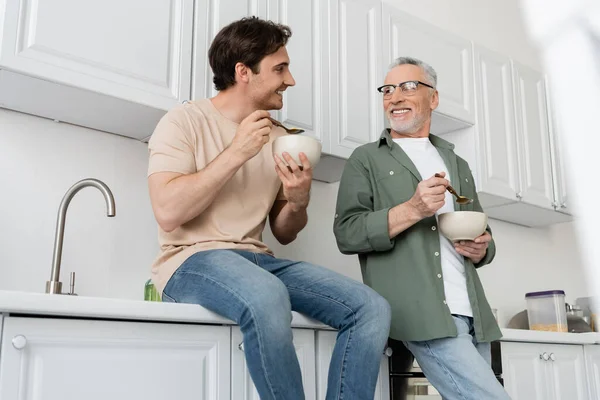 Smiling guy sitting on kitchen worktop and looking at happy dad during breakfast — Stock Photo