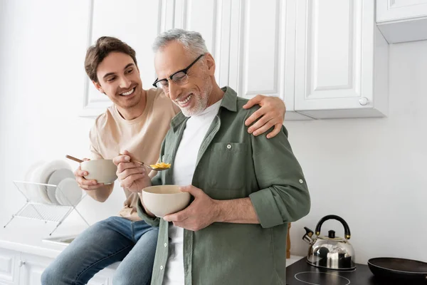 Happy young man sitting on kitchen worktop and hugging shoulders of smiling dad holding bowl and spoon with corn flakes — Stock Photo