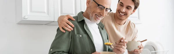 Cheerful guy hugging shoulders of smiling dad holding spoon with corn flakes during breakfast, banner — Stock Photo