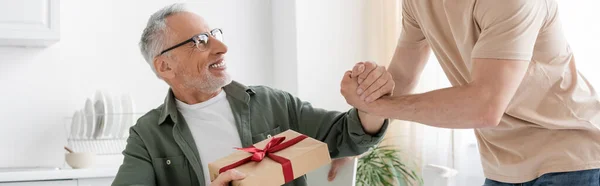 Улыбающийся мужчина держит подарочную коробку и пожимает руку сыну поздравляя его с Днем отца, баннер — Stock Photo