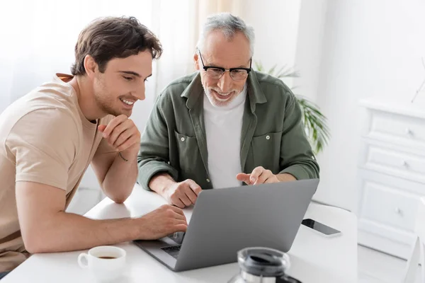 Happy mature man with smiling son looking at laptop near coffee pot in kitchen — Stock Photo