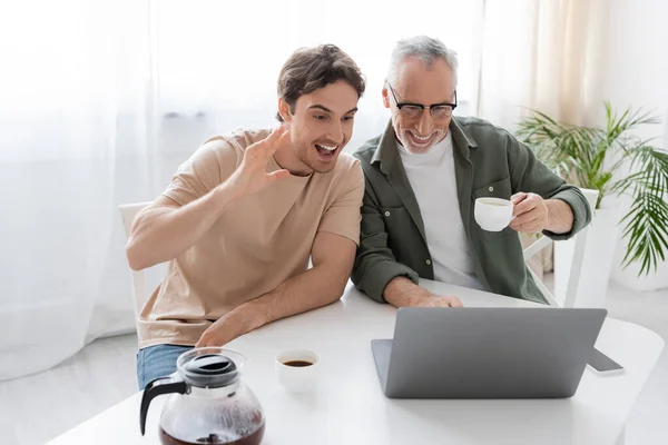 Excité homme agitant la main lors d'un appel vidéo sur ordinateur portable près père gai avec tasse de café — Photo de stock
