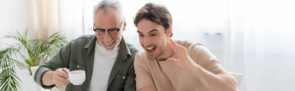 Jovem animado cara acenando mão perto sorrindo pai segurando xícara de café, banner — Fotografia de Stock