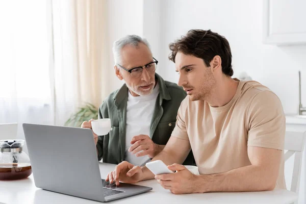 Grey haired man in eyeglasses holding coffee cup and pointing at laptop near adult son — Stock Photo
