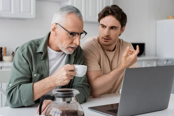 Jeune homme gestuelle tout en parlant à papa mature boire du café et en regardant ordinateur portable dans la cuisine — Photo de stock