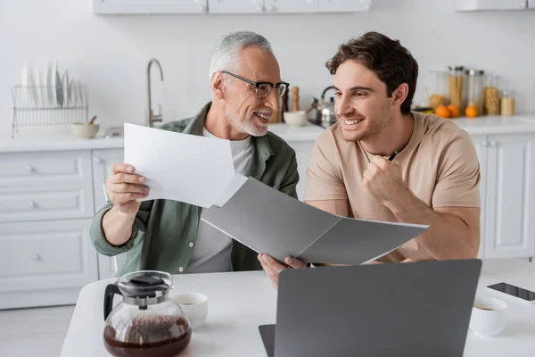 Smiling mature man holding documents near excited son showing win gesture near laptop and coffee pot — Stock Photo