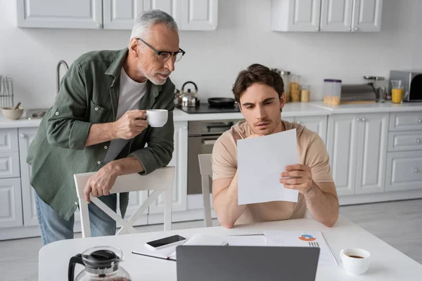 Sérieux jeune homme regardant document près de l'ordinateur portable et père aîné avec tasse de café — Photo de stock