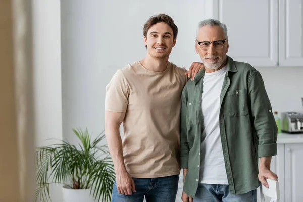 Happy young guy with grey haired father smiling at camera in kitchen — Stock Photo